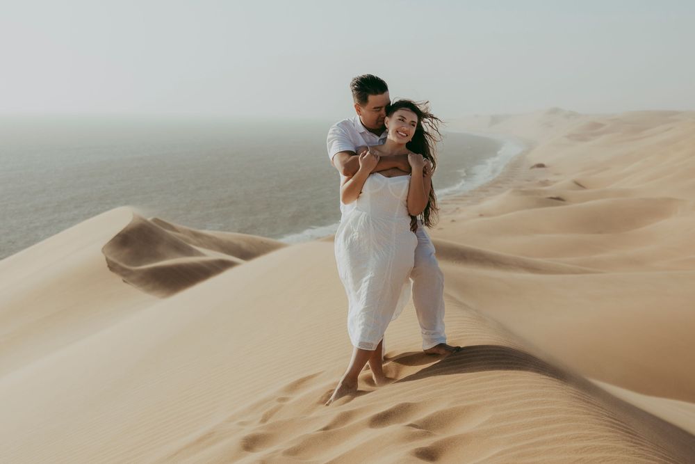 Newly engaged couple hugging at Sandwich Harbour, eyes closed, surrounded by soft sand and warm Namibian light.