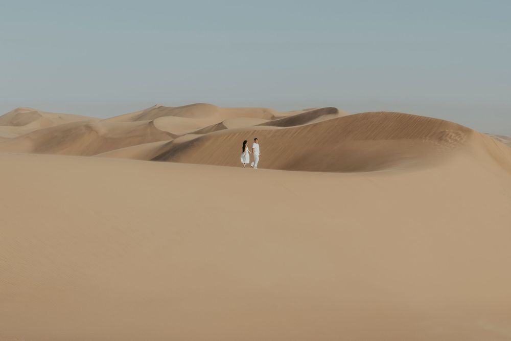 The couple walks barefoot along a soft sand ridge at Sandwich Harbour, Namibia, their footprints trailing behind.