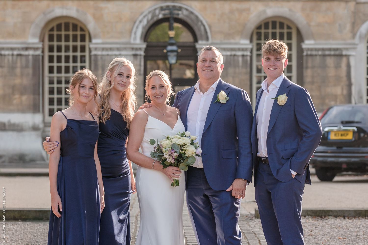 Wedding party members in navy blue and white attire pose together outside a grand stone building.