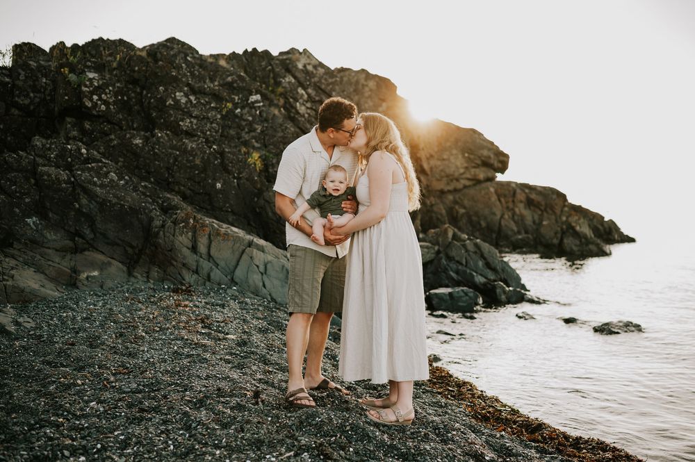 Family photo of parents holding baby kissing at Neck Point Park Nanaimo golden hour family photography by Vancouver Island photographer