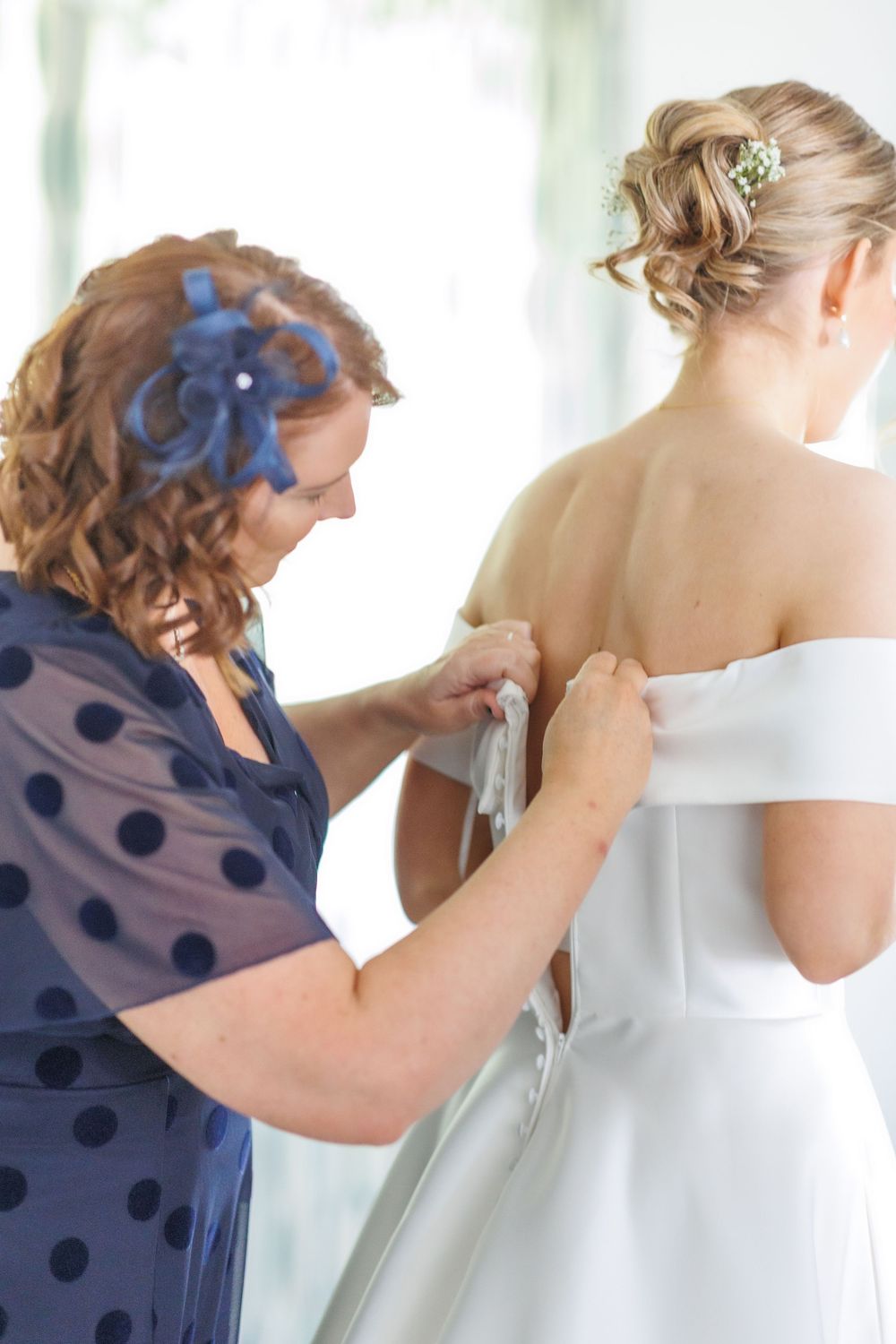 A bride in a white wedding dress is helped with final adjustments by someone wearing a navy polka dot dress.