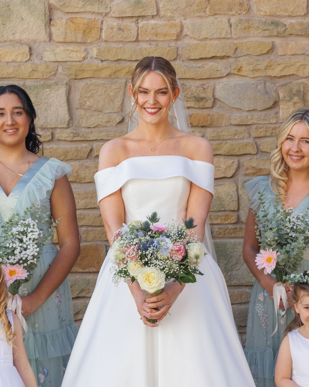 A bridal party stands together holding bouquets with white and blue flowers against a stone wall background.