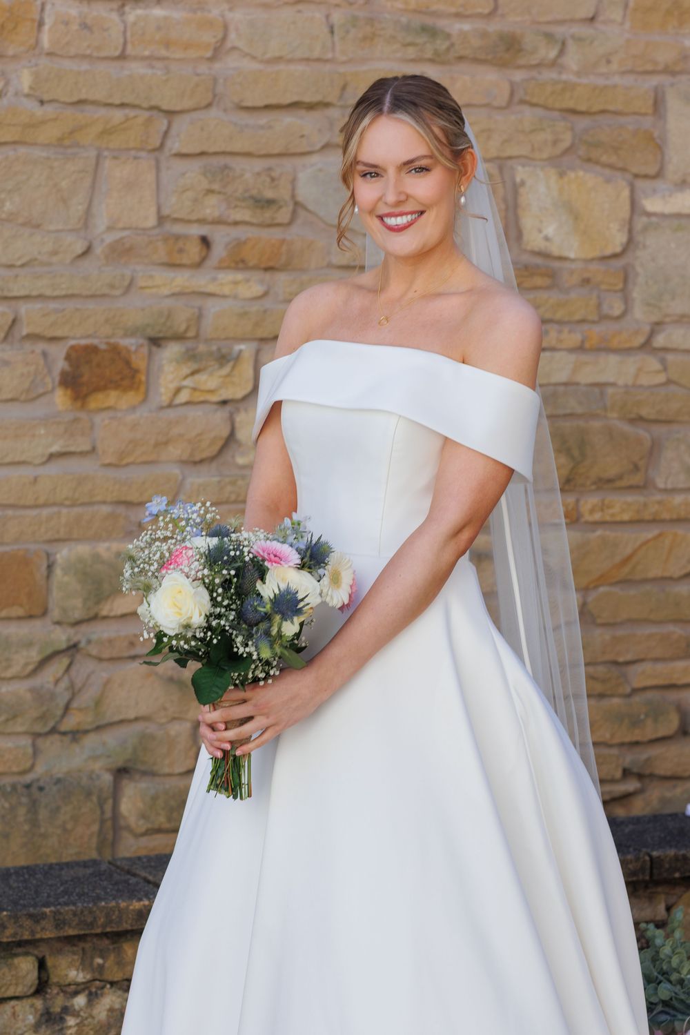 Elegant bride in off-shoulder white wedding dress holding floral bouquet against stone wall.