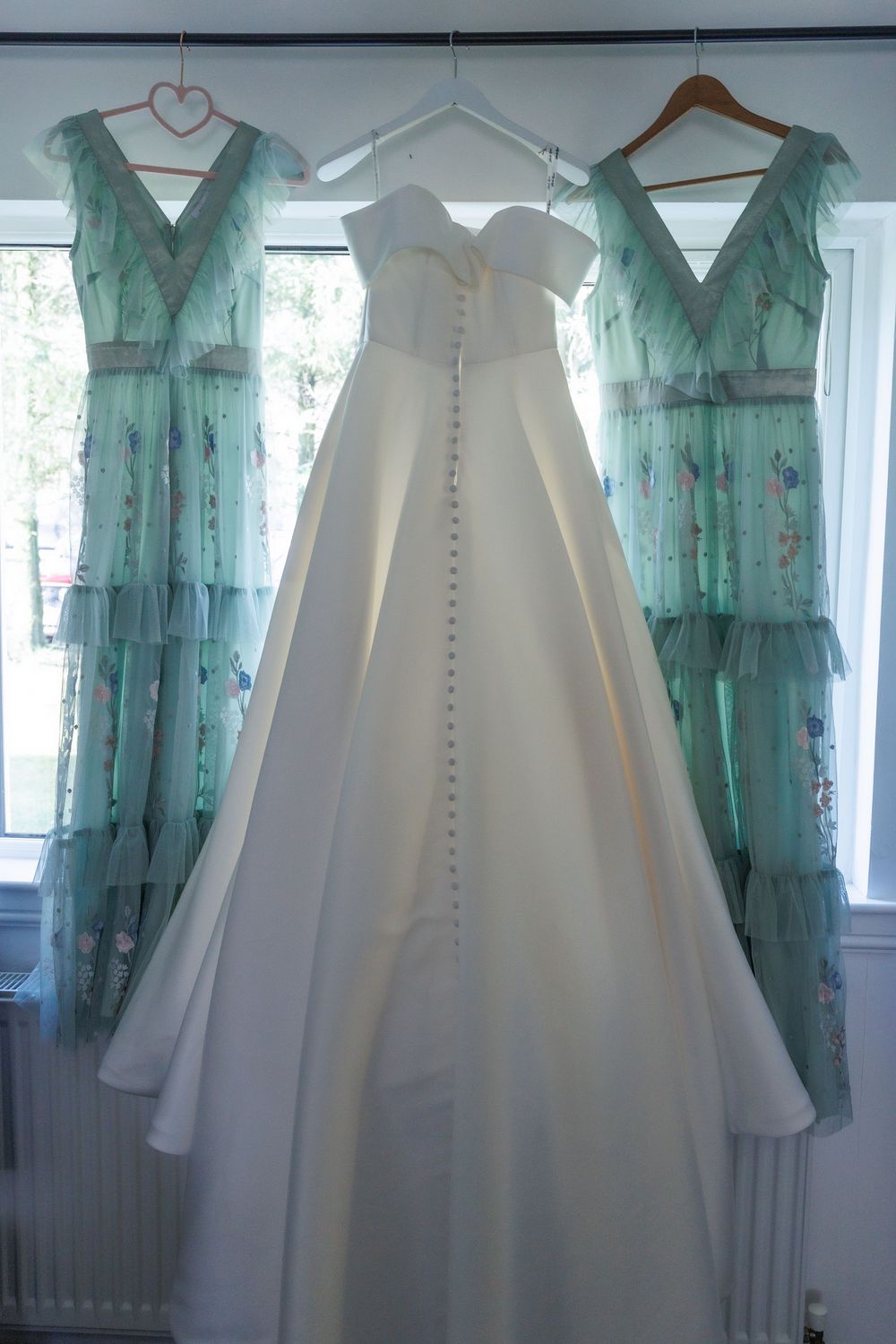Wedding gown and two mint green bridesmaid dresses hanging on display against a window.