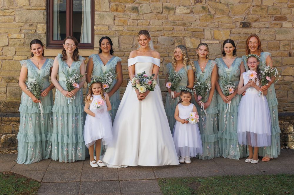 Wedding party wearing sage green bridesmaid dresses and white flower girl dresses pose together against stone wall.