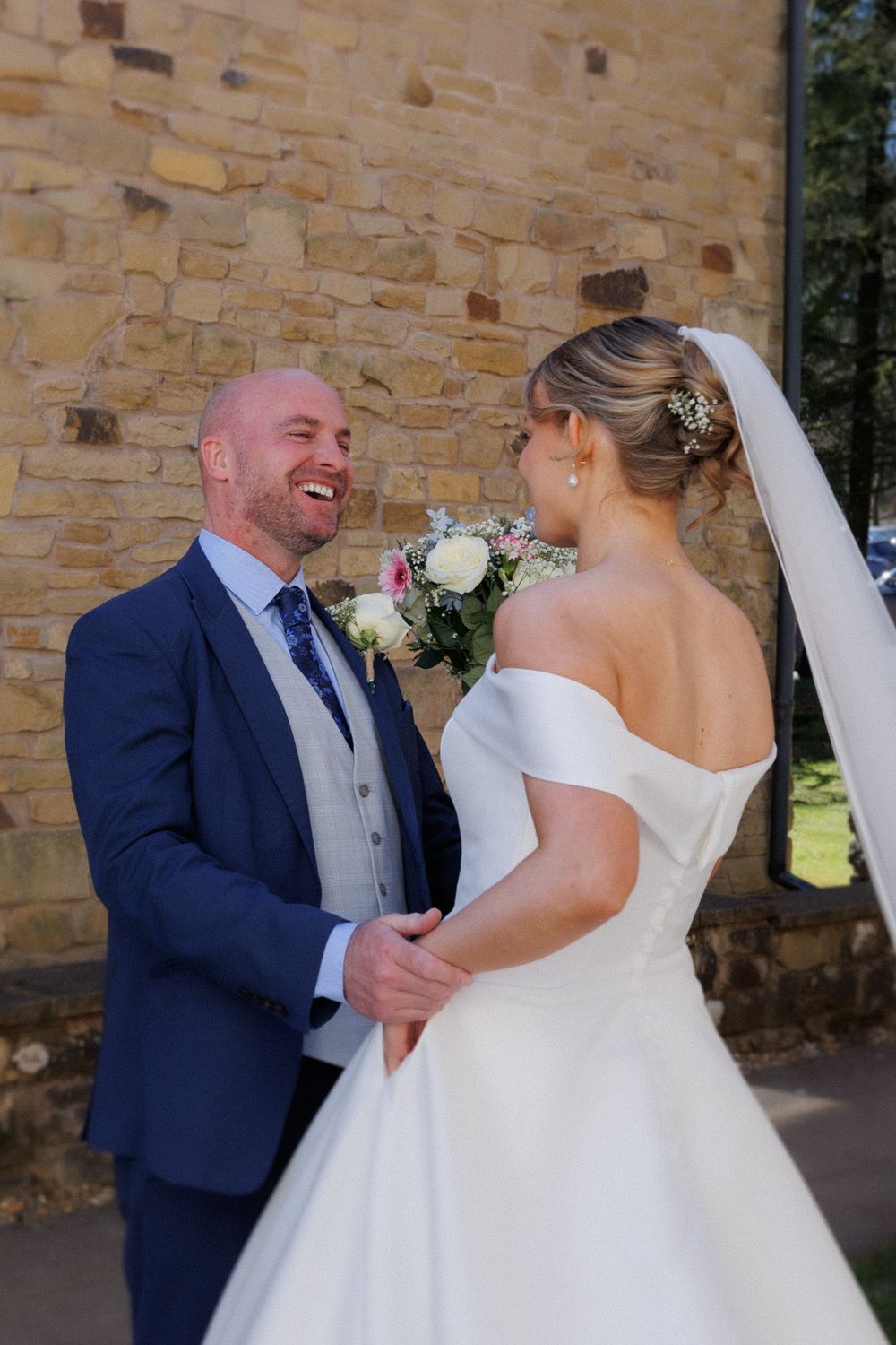 A romantic moment between newlyweds sharing a joyful laugh against a rustic stone wall backdrop on their wedding day.