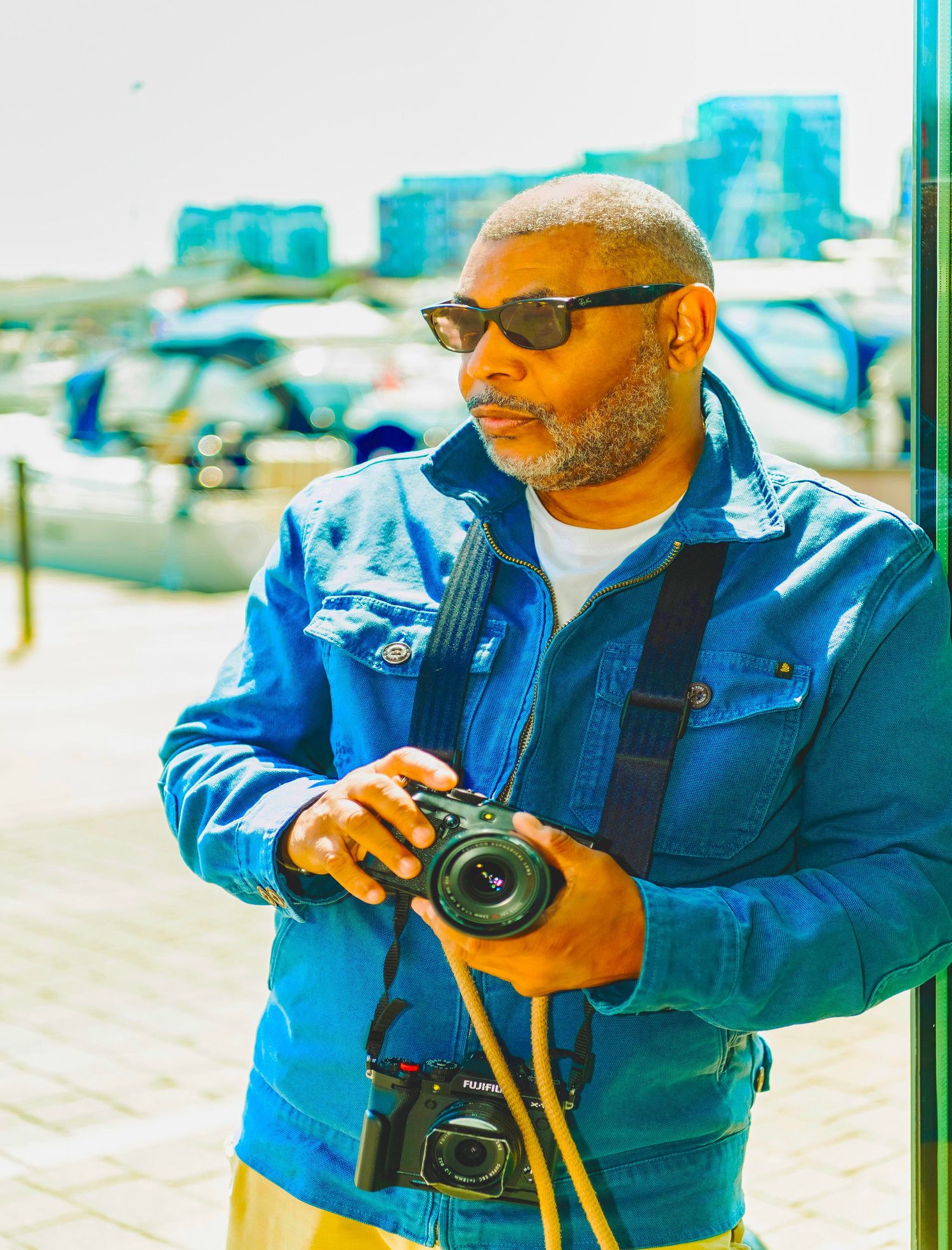 A photographer in sunglasses and blue denim jacket holds a digital camera while standing outdoors on a sunny day.
