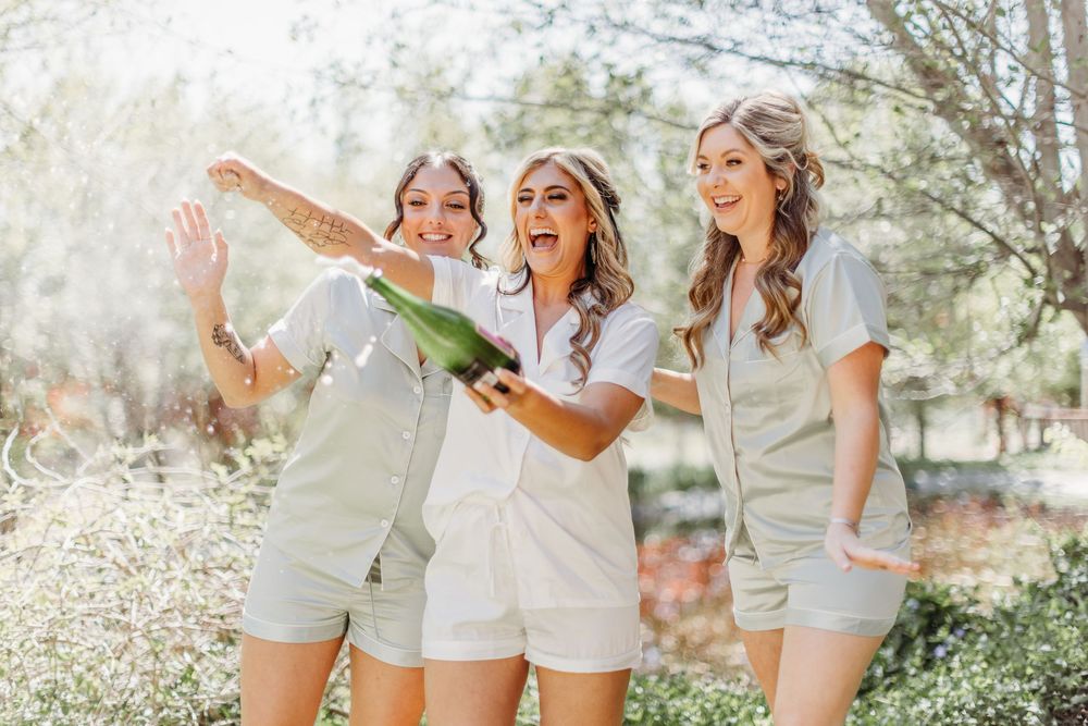 Three women in matching outfits celebrating with champagne in a sun-drenched garden.