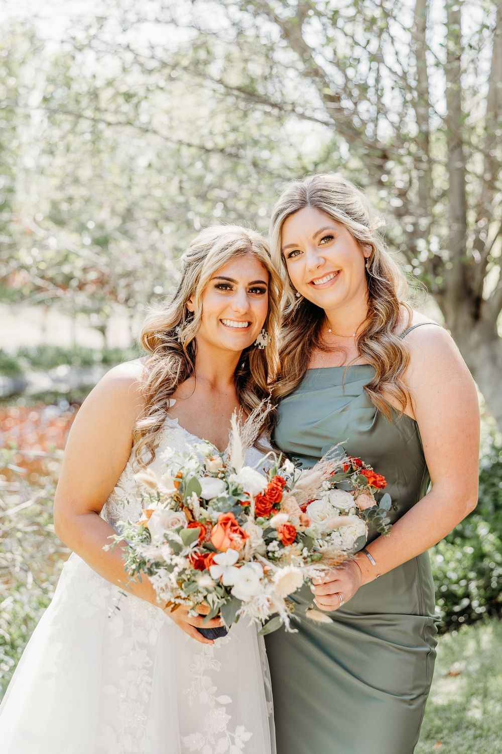 Two women pose outdoors holding floral bouquets, one in a white wedding dress and one in a sage green dress.