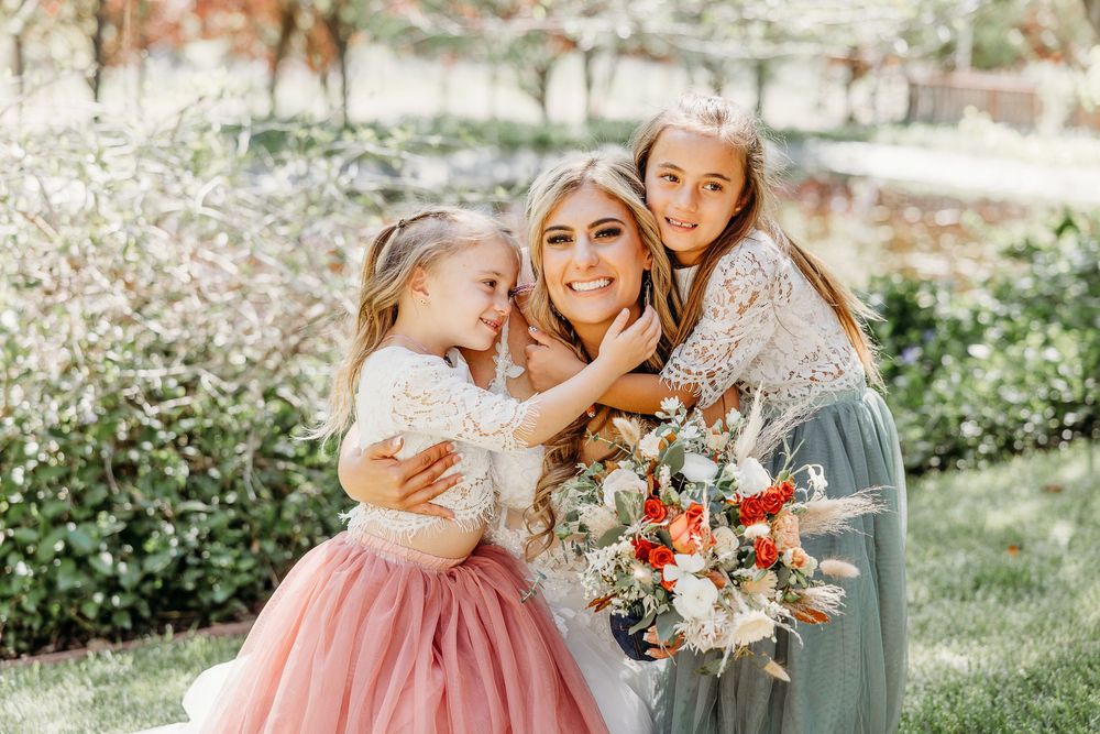 A bride in a wedding dress sharing a tender moment with flower girls in the garden.
