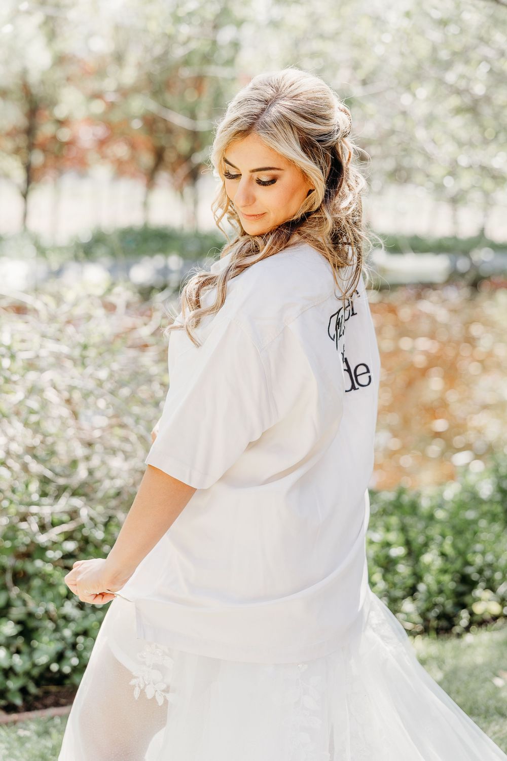 A bride in a white robe poses elegantly in a sun-dappled garden setting.
