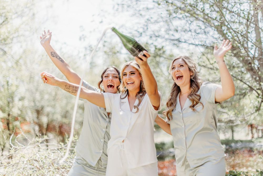 Three bridesmaids in light colored dresses celebrating joyfully with champagne outdoors.