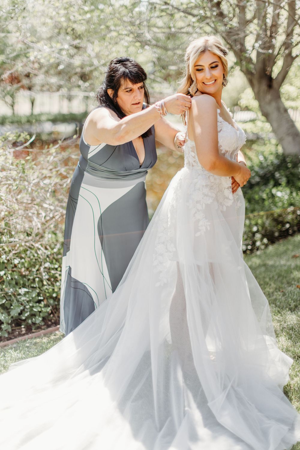 A bride in a white lace gown gets final adjustments made to her dress outdoors in a garden setting.
