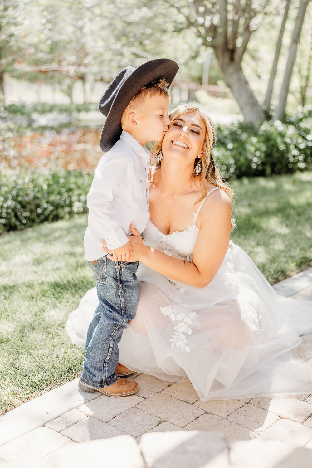 A young boy in a cowboy hat shares a sweet moment with the bride on her wedding day in a garden.