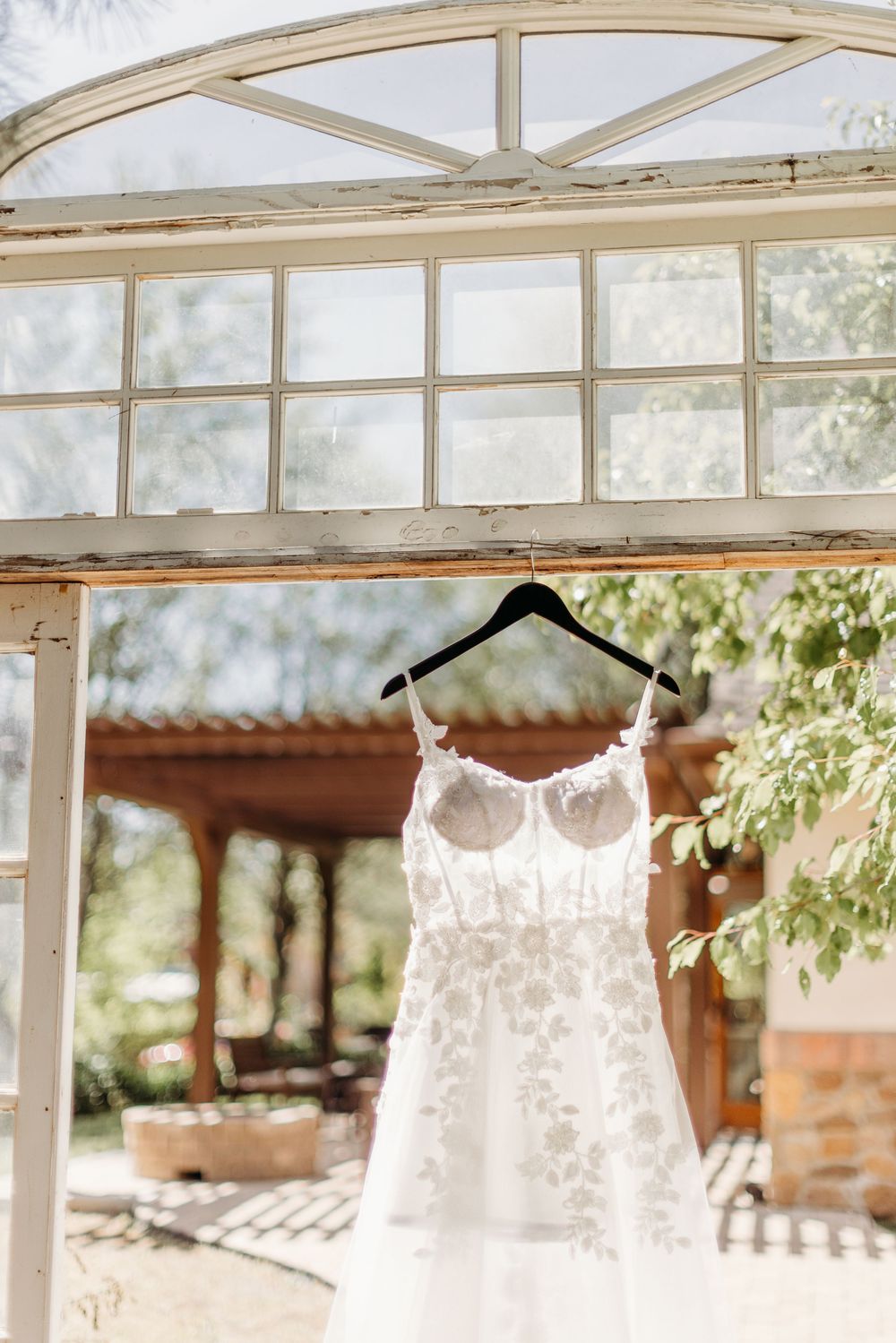 A delicate white lace wedding dress hanging from a hanger against an arched window frame in natural light.