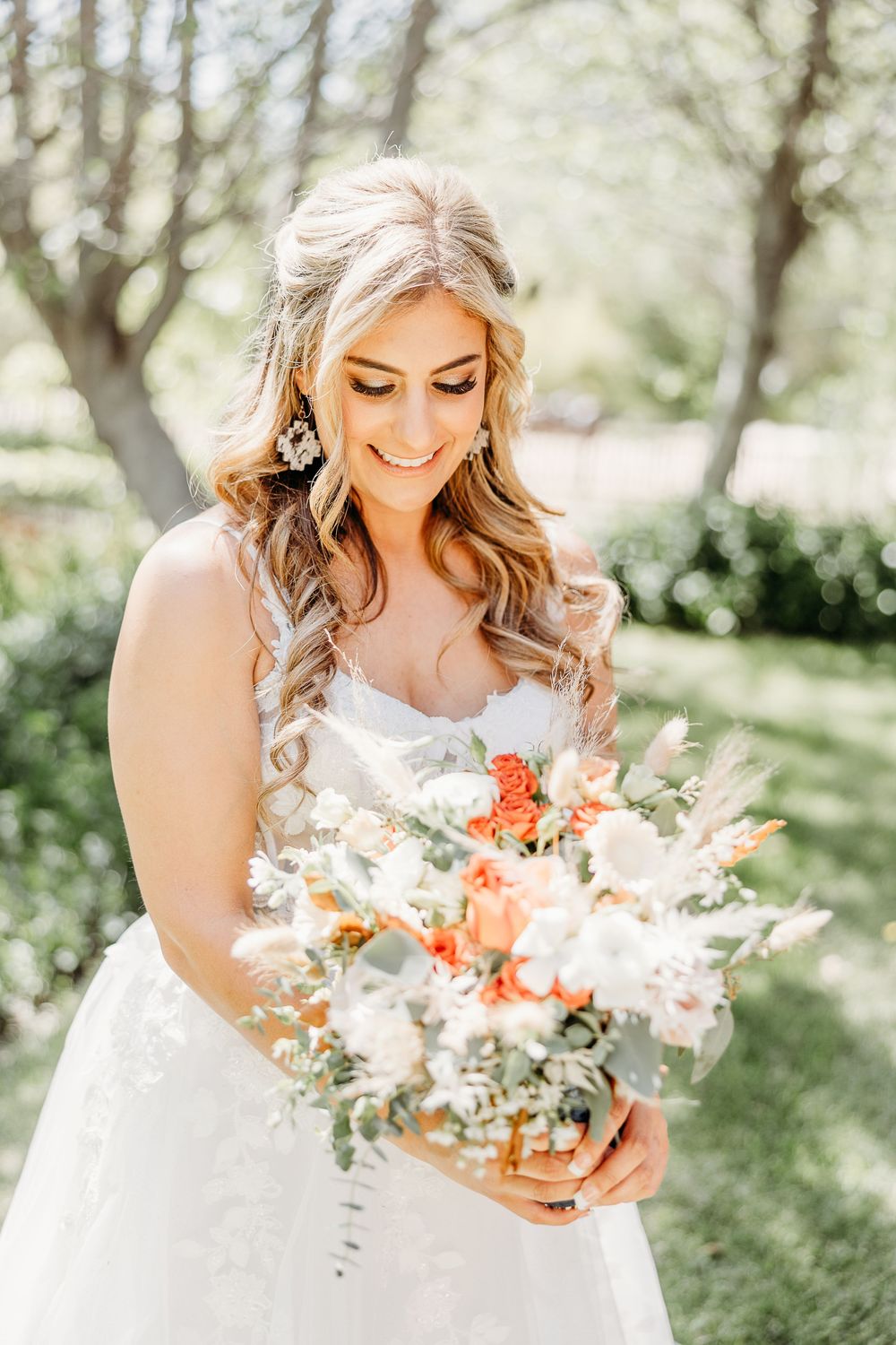 A bride smiles while holding a beautiful bouquet of coral and white flowers in a sunny garden setting.
