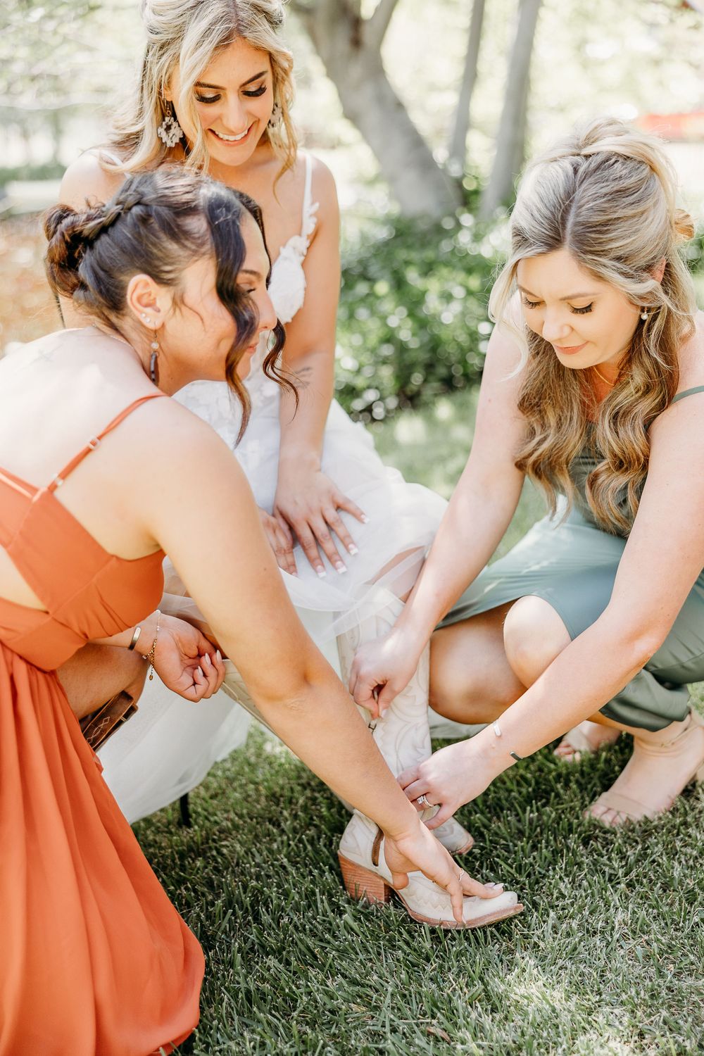 Bridesmaids in coral dresses help adjust the bride's shoes on a grassy lawn.