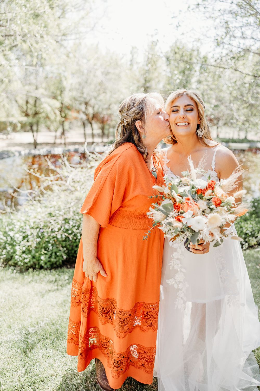 A tender moment between a bride in white and a woman in an orange dress holding a floral bouquet.