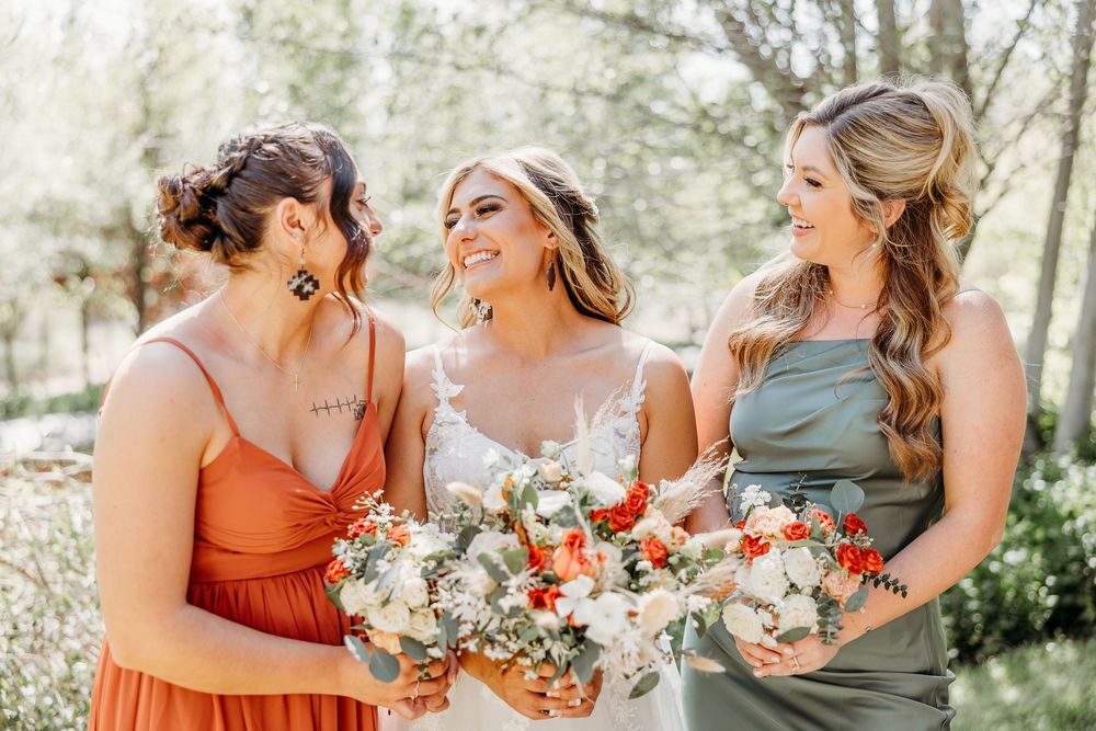 Bridesmaids in orange and sage dresses laughing while holding floral bouquets.
