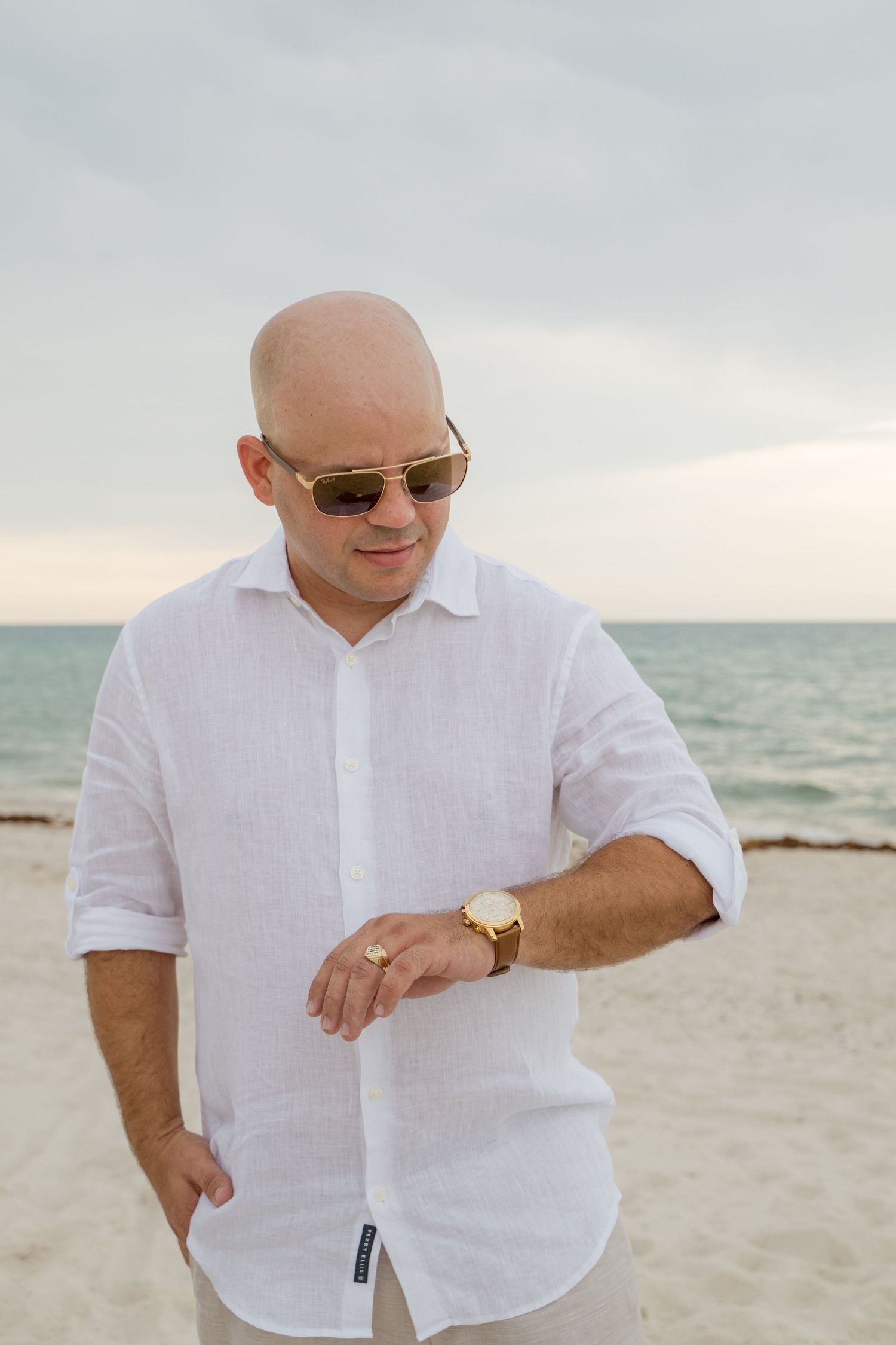 A person in a white linen shirt and sunglasses checks their gold watch while standing on a beach near the ocean at dusk.