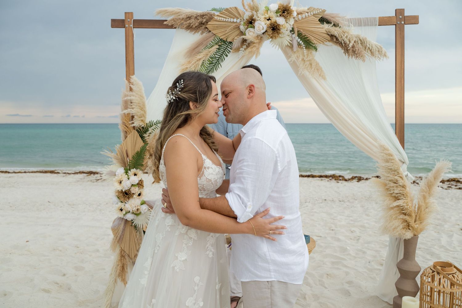 A romantic beach wedding setup with wooden crosses, pampas grass, and floral arrangements at sunset.