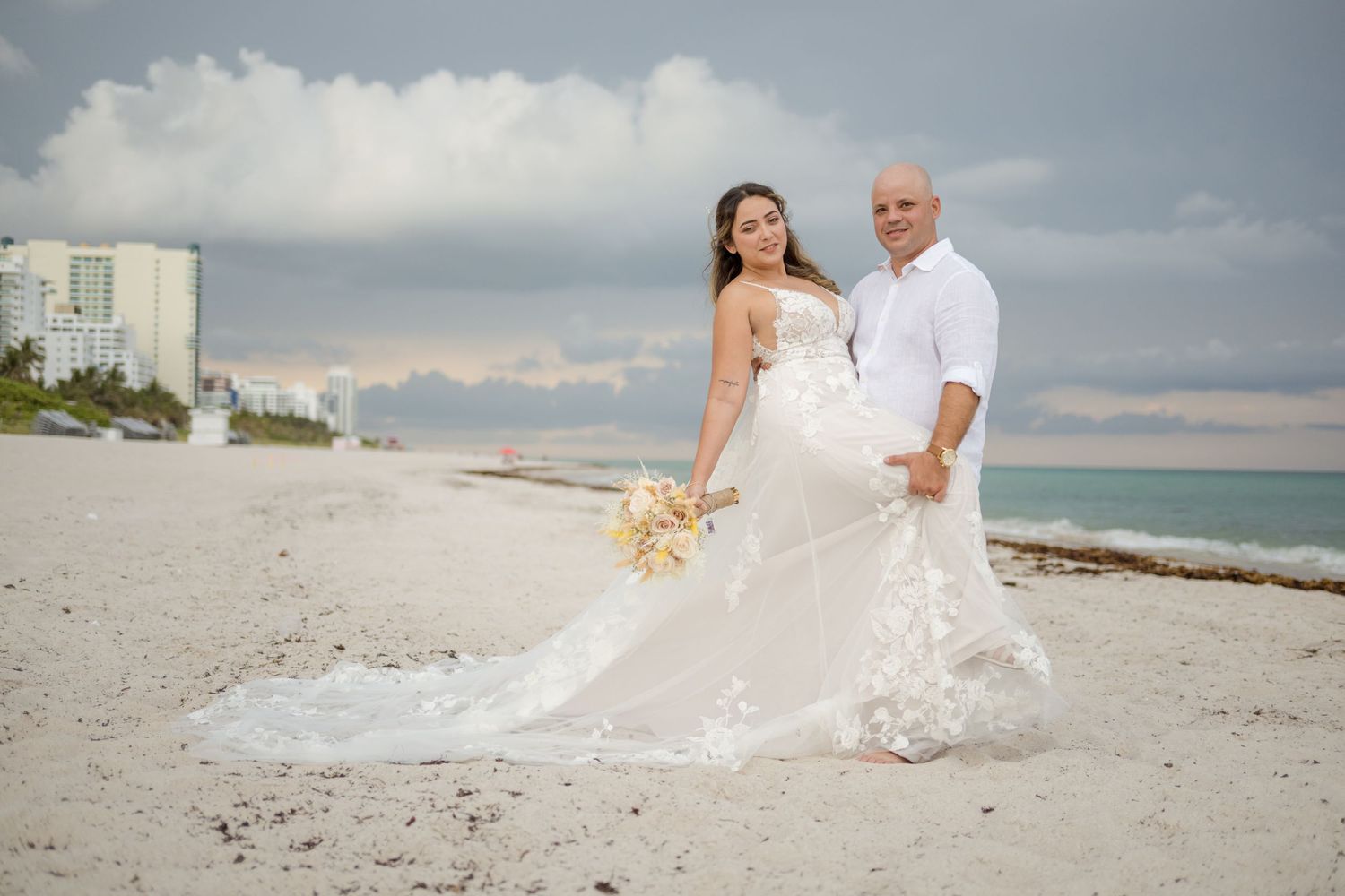 Newlywed couple poses for romantic beach wedding photo with white dress flowing in ocean breeze at sunset.
