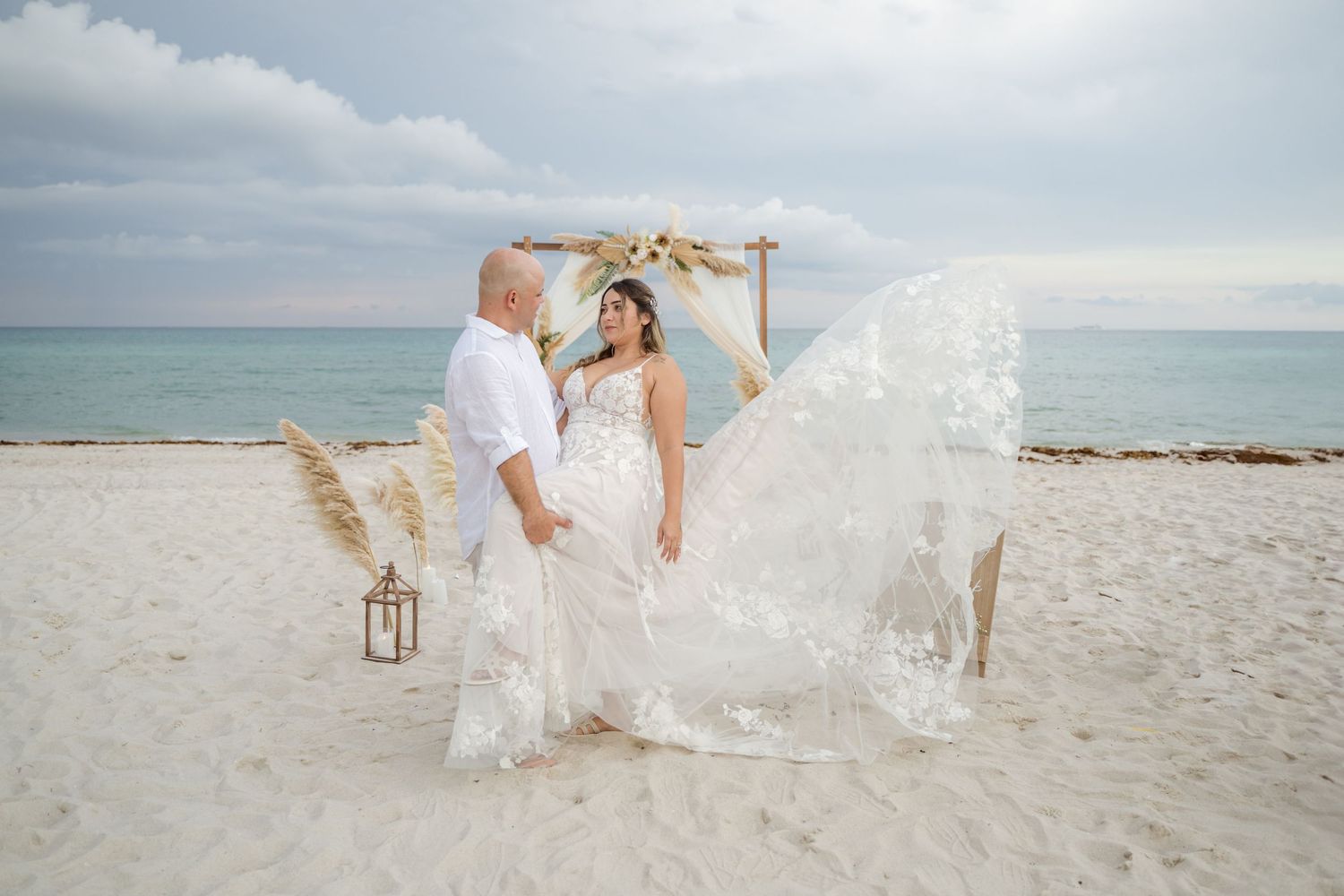 A beach celebration scene with flowing white fabric and pampas grass decorations against the ocean backdrop.