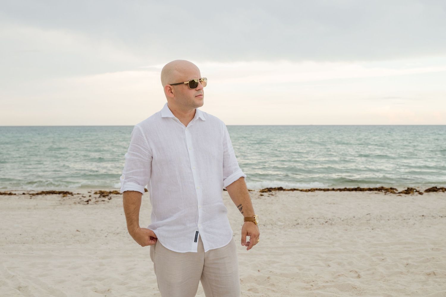 A person in a white button-down shirt and khaki pants stands on a peaceful beach at sunset with ocean waves in background.
