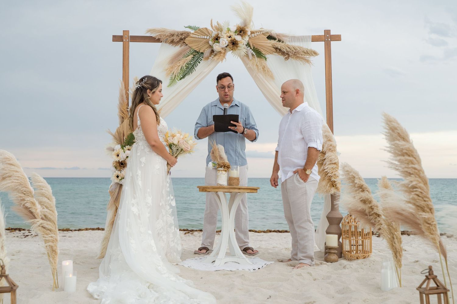 Beach wedding ceremony setup with pampas grass arch and couple exchanging vows by the ocean.