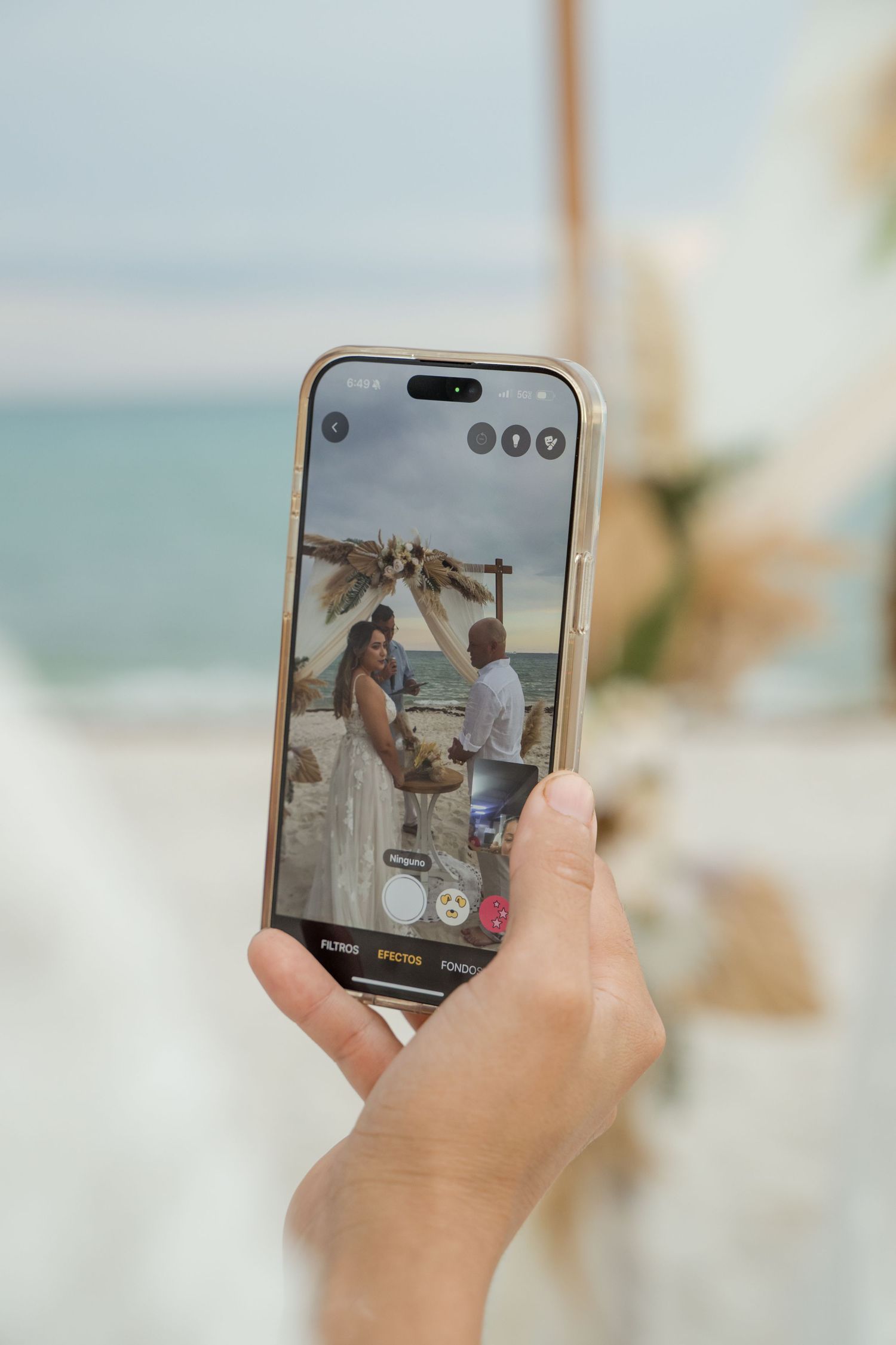 A hand holds a smartphone showing a beach wedding ceremony being photographed through the phone's camera screen.