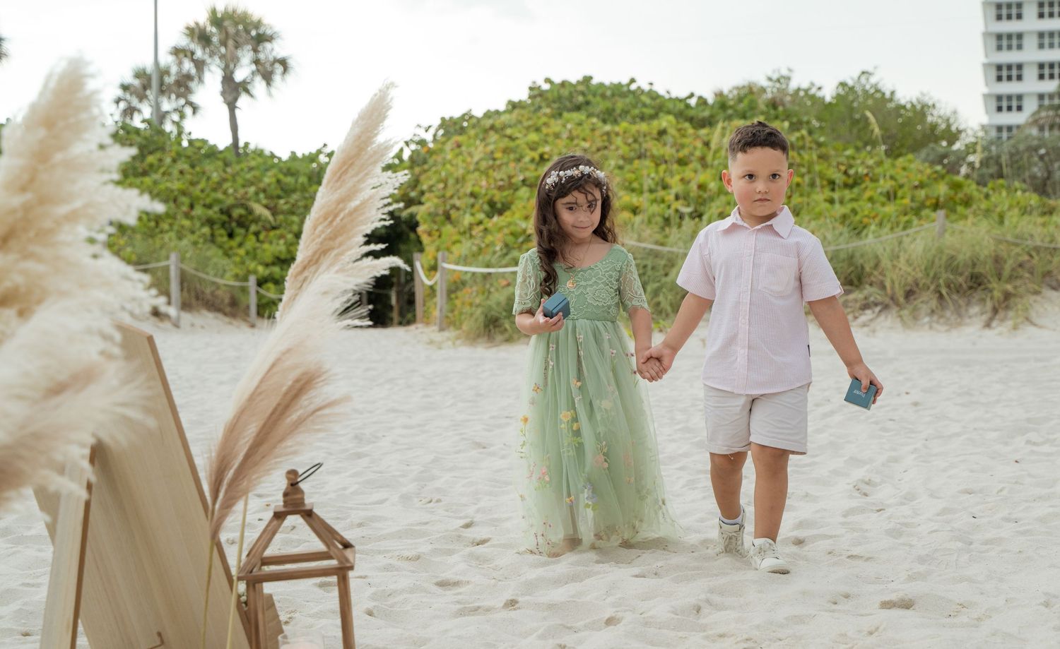 Two children hold hands while walking on a sandy beach with pampas grass in the foreground and palm trees behind them.