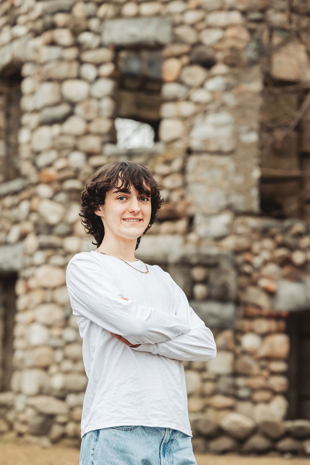 A person in a white long-sleeve shirt and jeans stands with arms crossed against a rustic stone wall background.