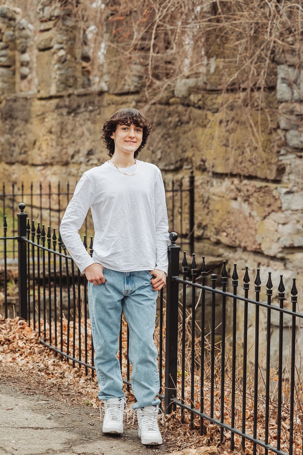 A person in a white long sleeve shirt and light jeans leans casually against a wrought iron fence near a stone wall.