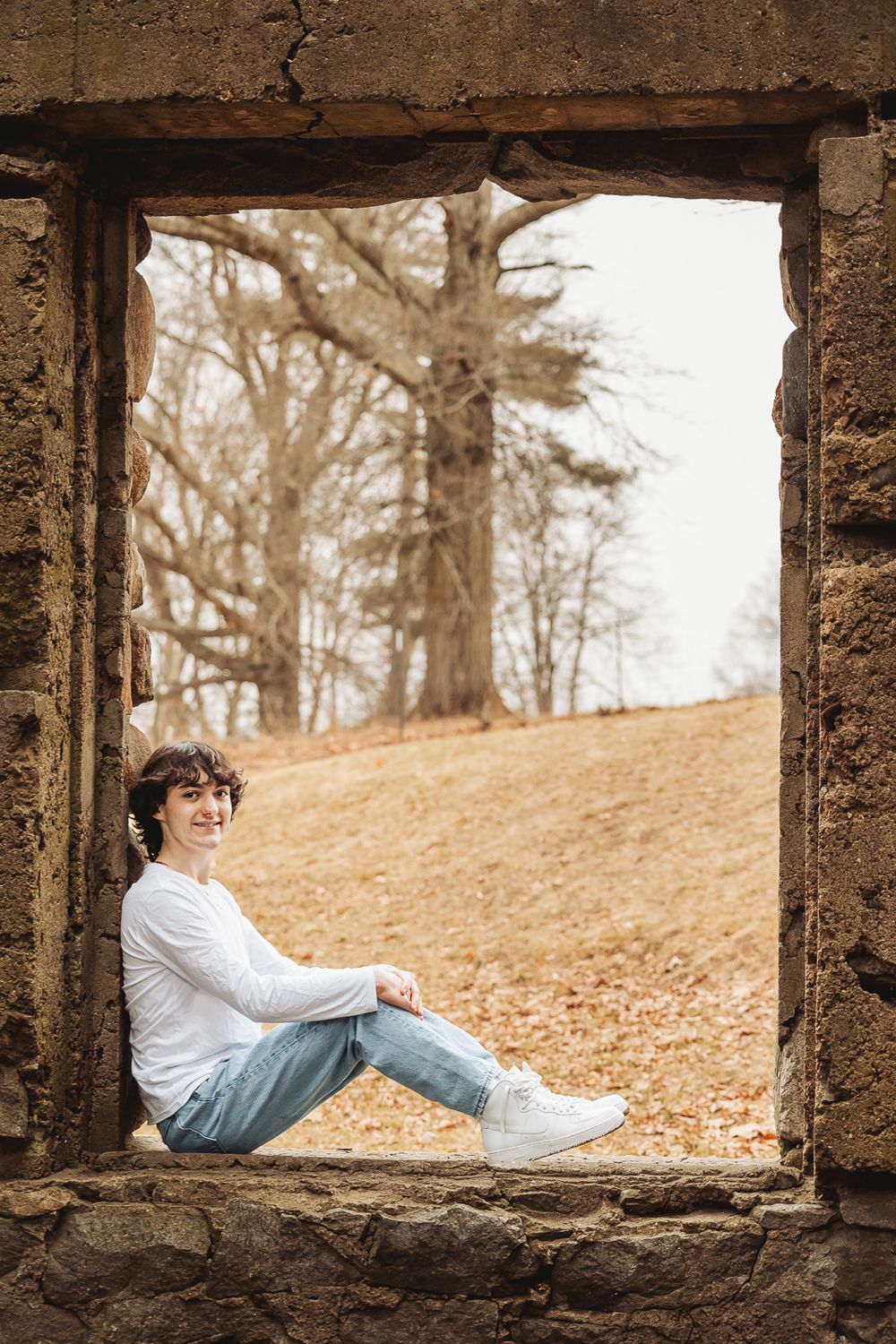 Person sitting in a stone window frame surrounded by autumn leaves and bare trees in a natural setting.