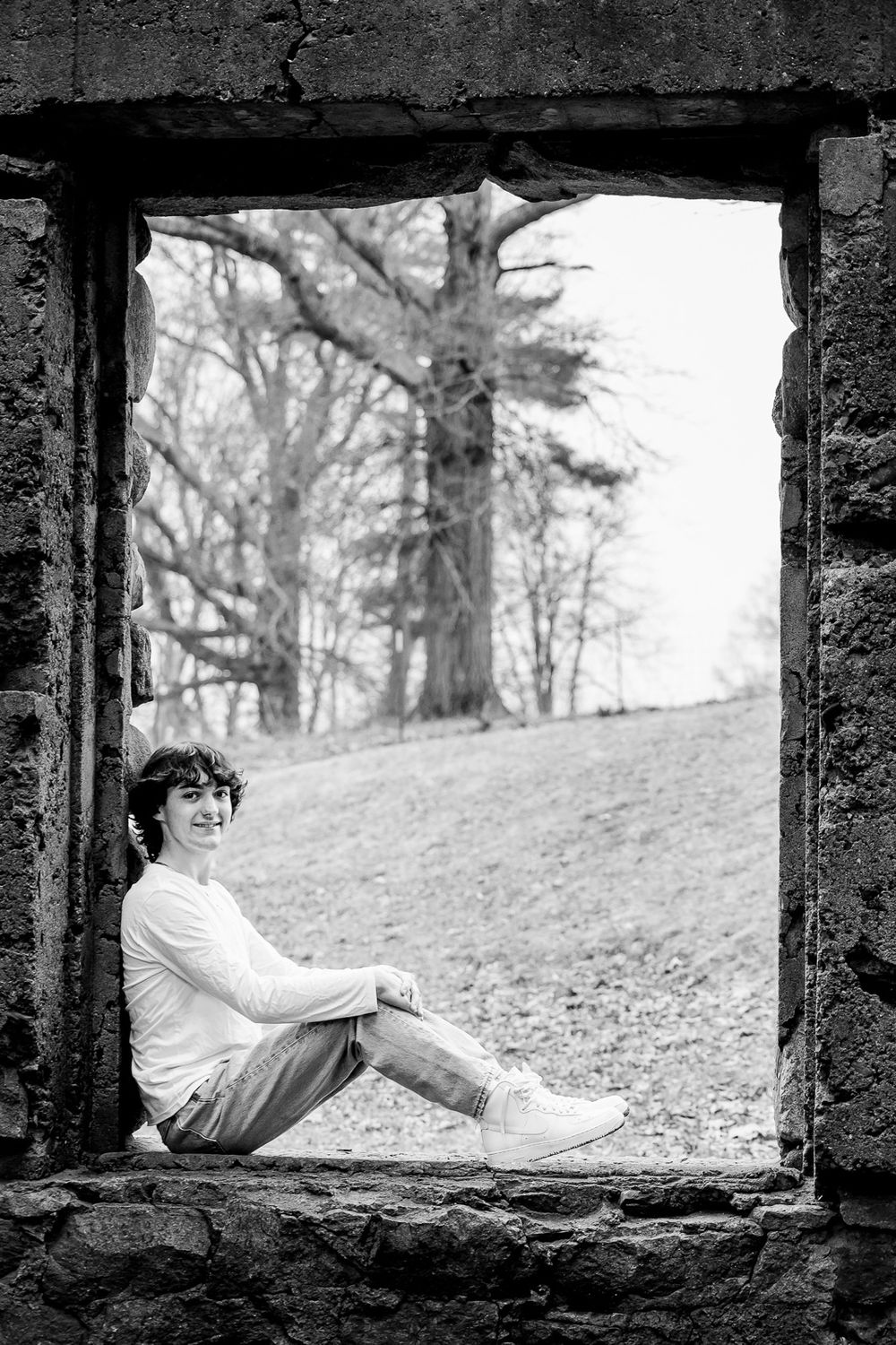 A figure relaxes in a dramatic horizontal pose, framed through a stone or concrete opening in black and white.