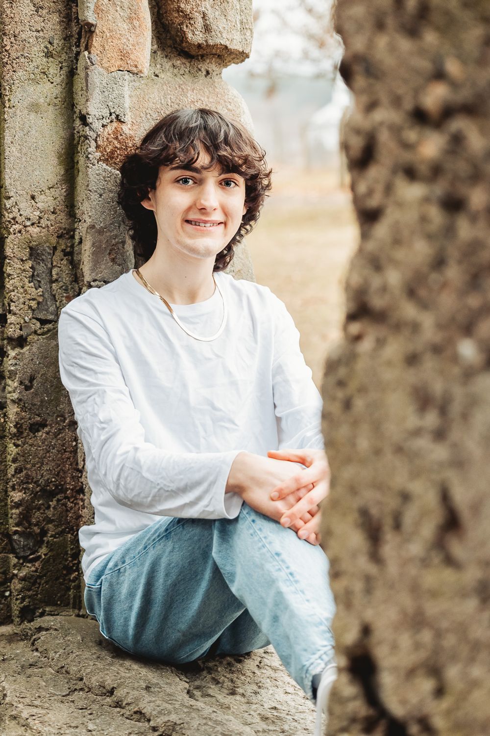 A person in a white sweater and jeans sits casually against an old stone wall in an outdoor setting.