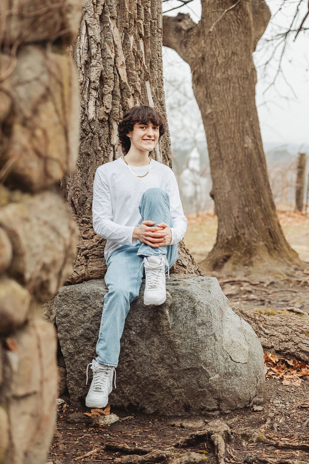 A person in a white sweater and light jeans sits casually on a large rock by tree trunks in an autumn forest setting.