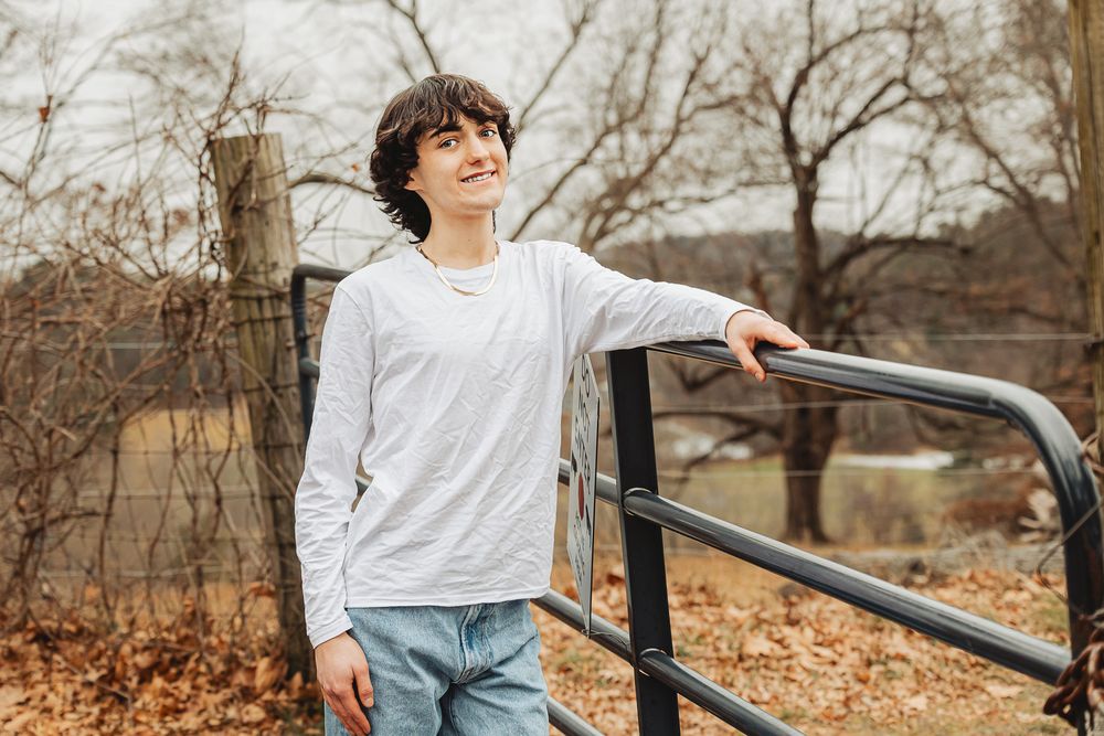 A person in a white long sleeve shirt leans against a metal railing surrounded by autumn foliage in a natural setting.