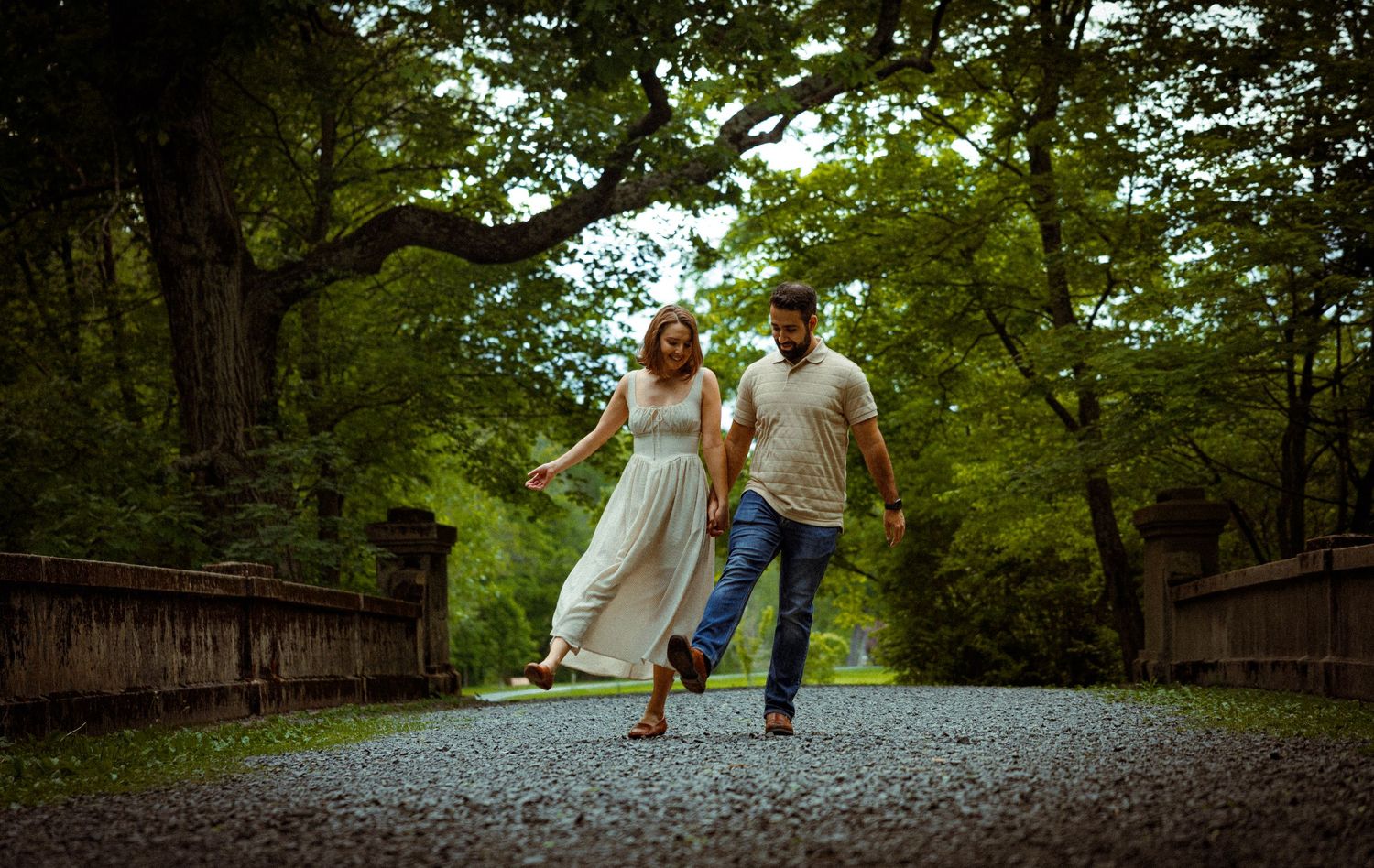 A couple holds hands while walking together on a tree-lined path in a lush green park on a sunny day.