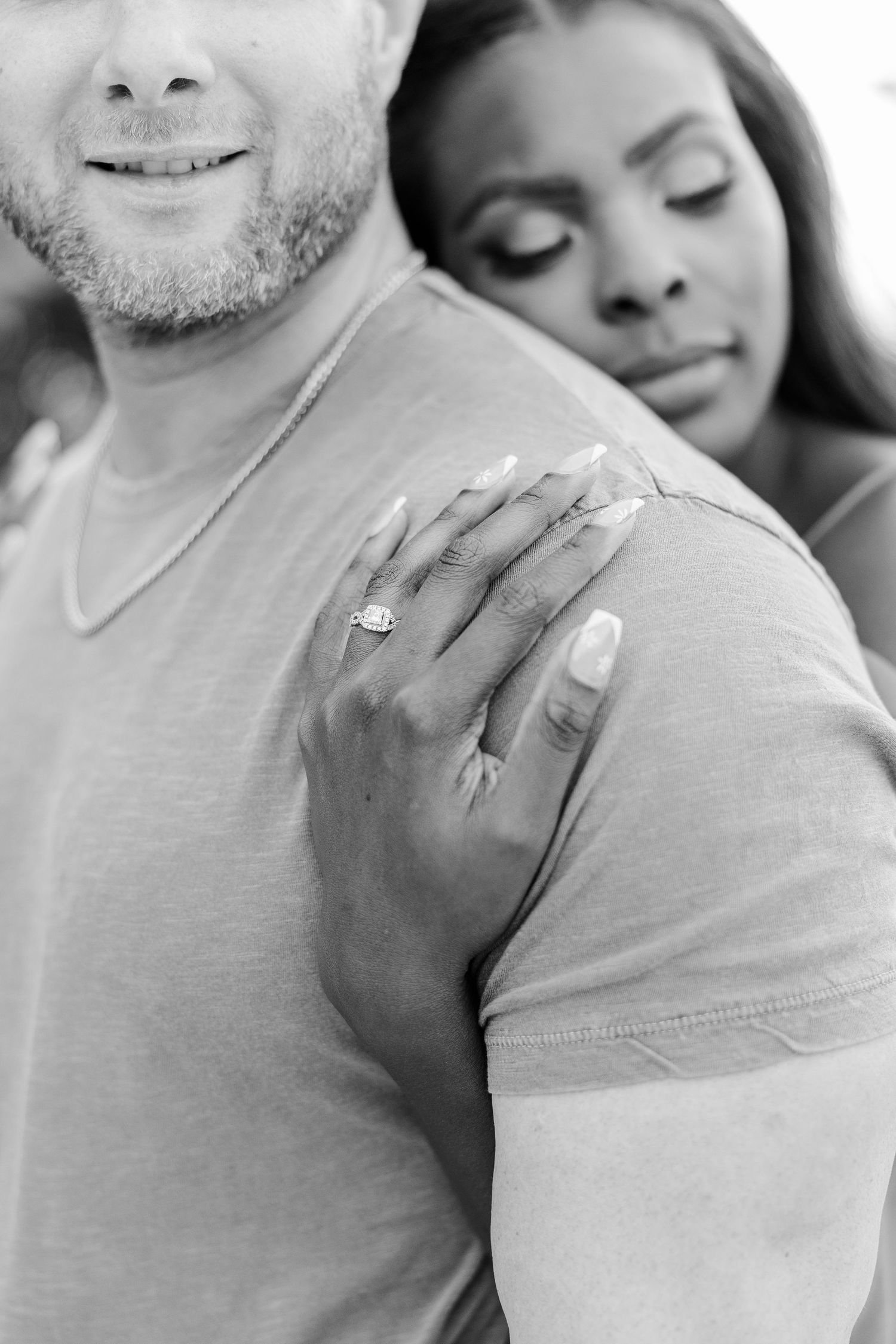 Close-up black and white photo showing a hand with an engagement ring resting on a shoulder in an intimate embrace.