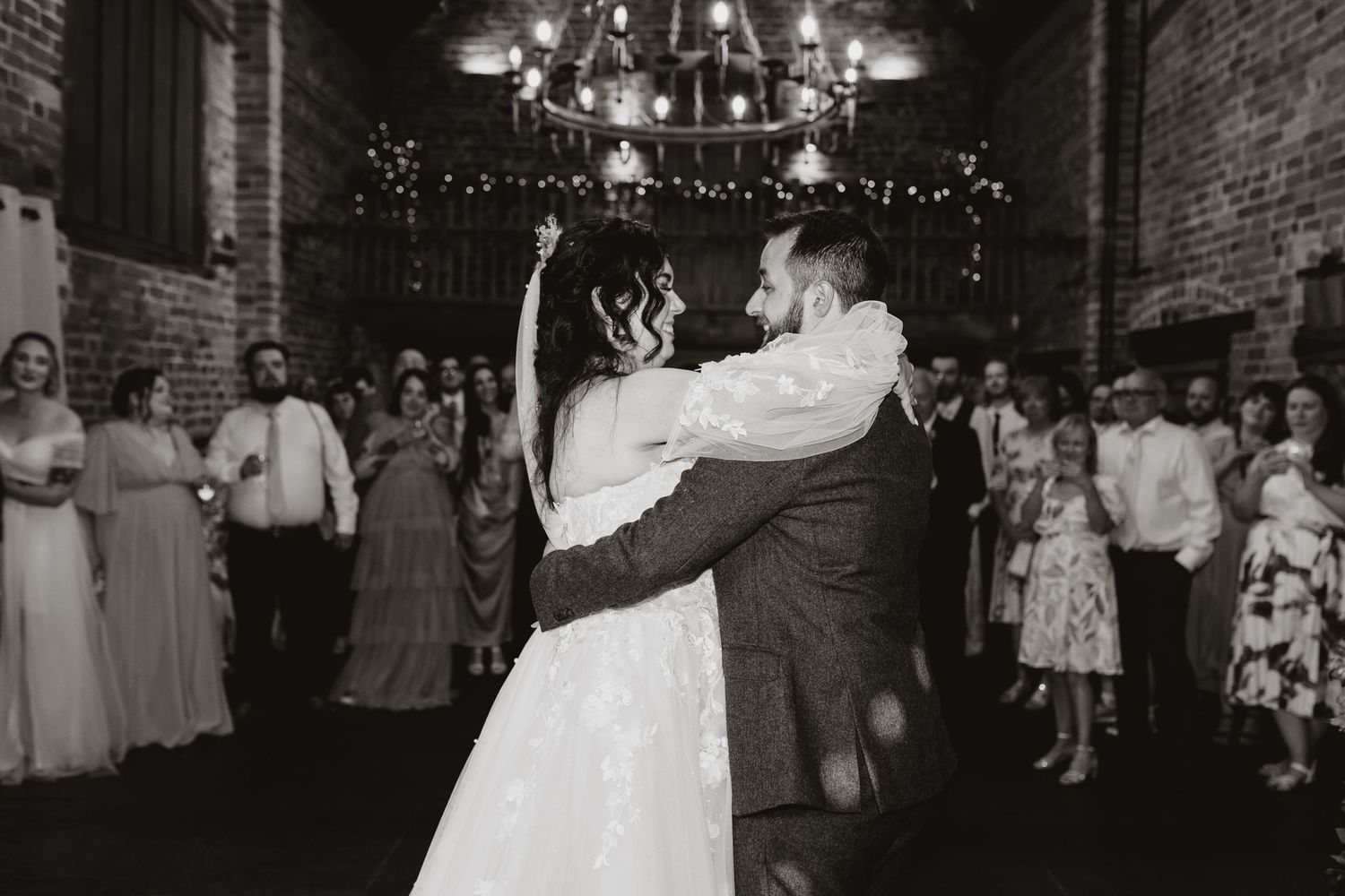 Couple shares their first dance at a rustic brick-walled reception venue decorated with twinkling lights and guests watching.