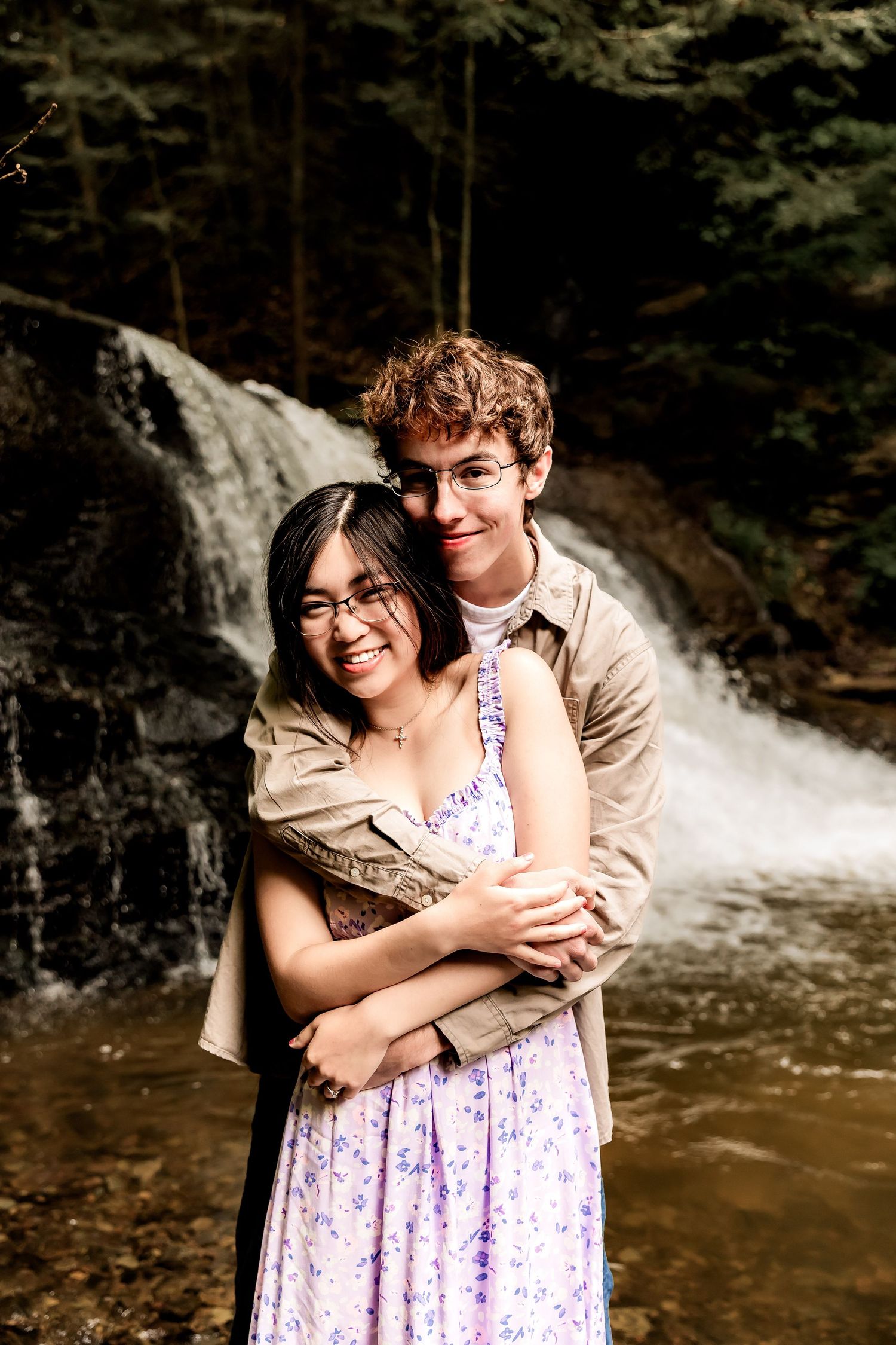 A couple embraces in front of a cascading waterfall in the forest, with her wearing a floral dress and him in beige.