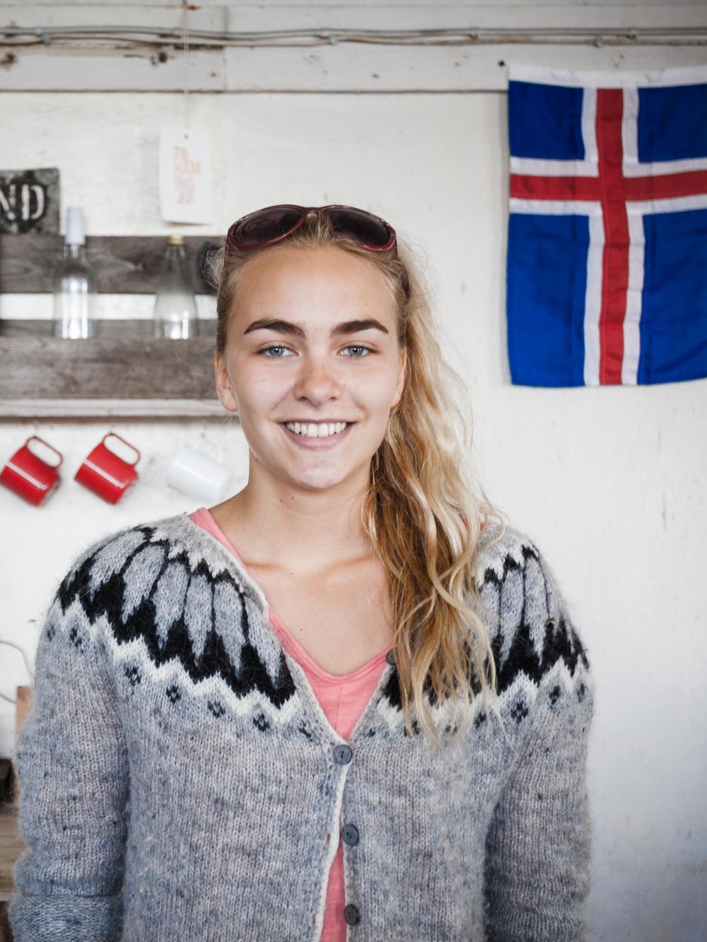 A blonde person wearing a traditional Icelandic sweater stands smiling in front of an Iceland flag on a white wall.