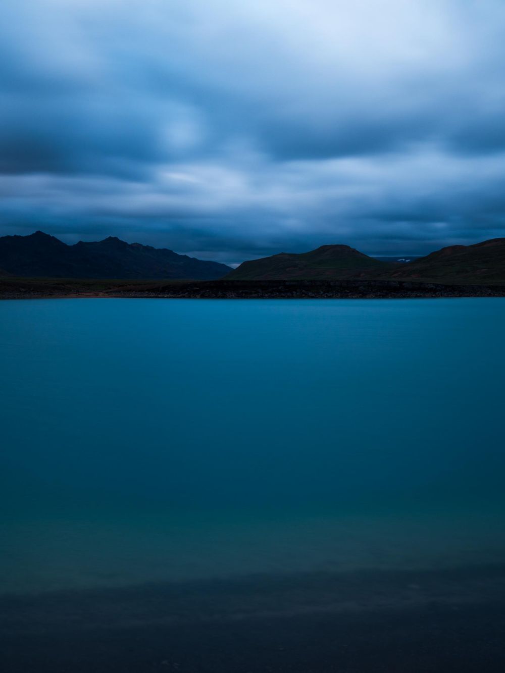 Dramatic turquoise lake reflecting moody storm clouds and dark mountain silhouettes at dusk.