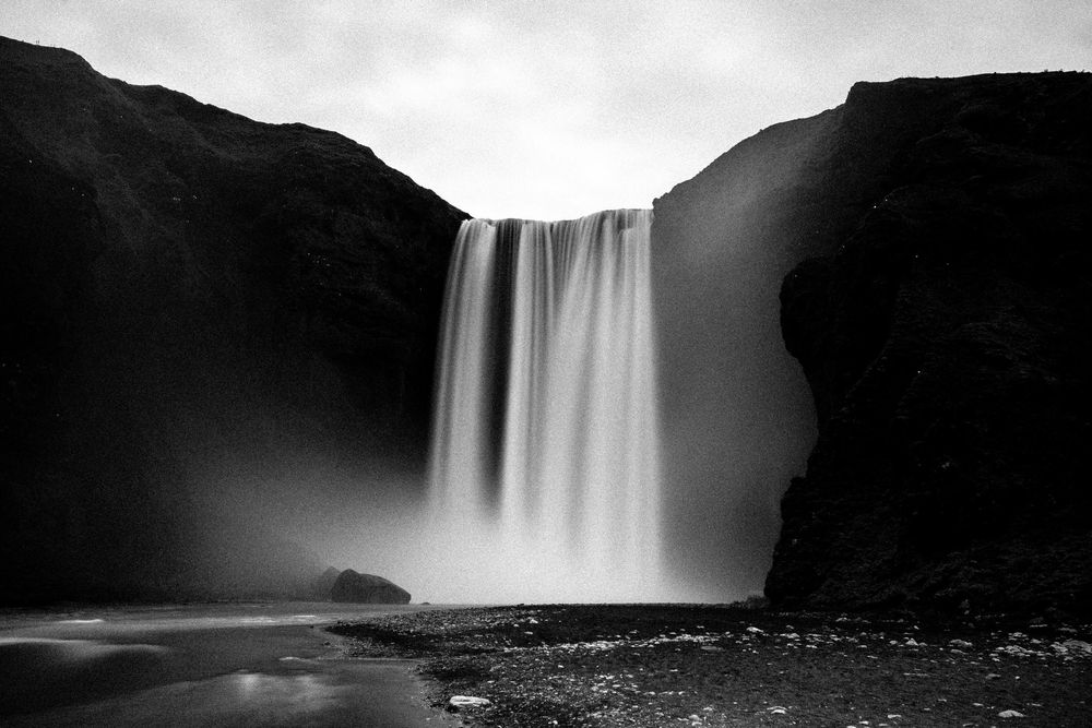 A dramatic black and white photograph of Skogafoss waterfall in Iceland cascading down towering cliffs into a misty pool below.