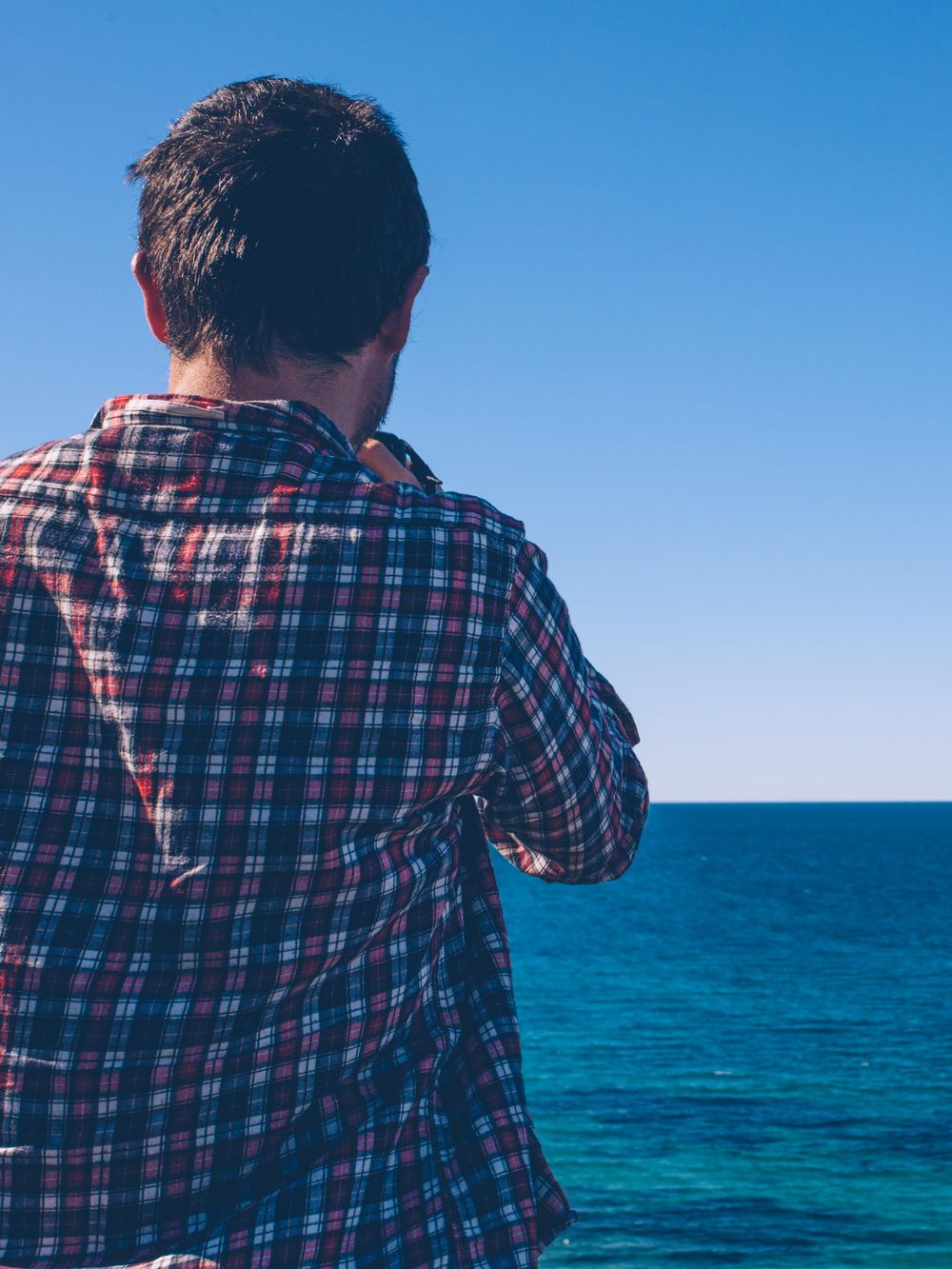Person in plaid shirt looking out at vibrant blue ocean waters against clear sky on sunny day.