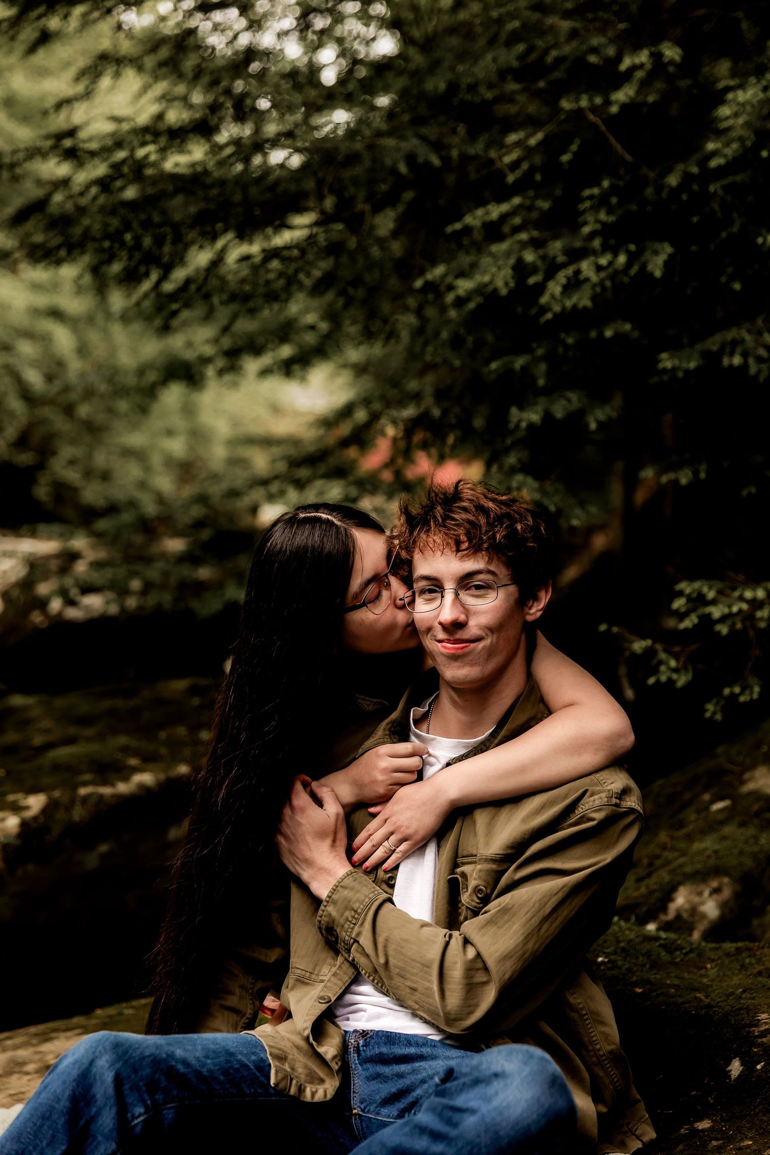 A couple shares an intimate moment outdoors in a wooded setting with soft natural lighting filtering through the trees.