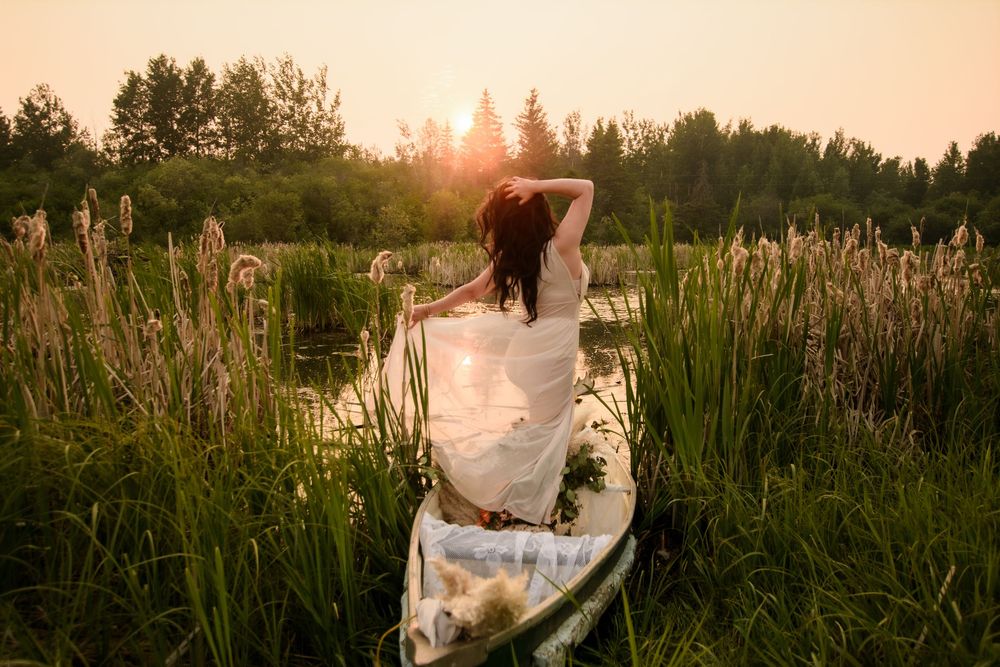 A dreamy sunset scene shows a woman in white dress sitting in a rustic boat surrounded by cattails and marsh grass.