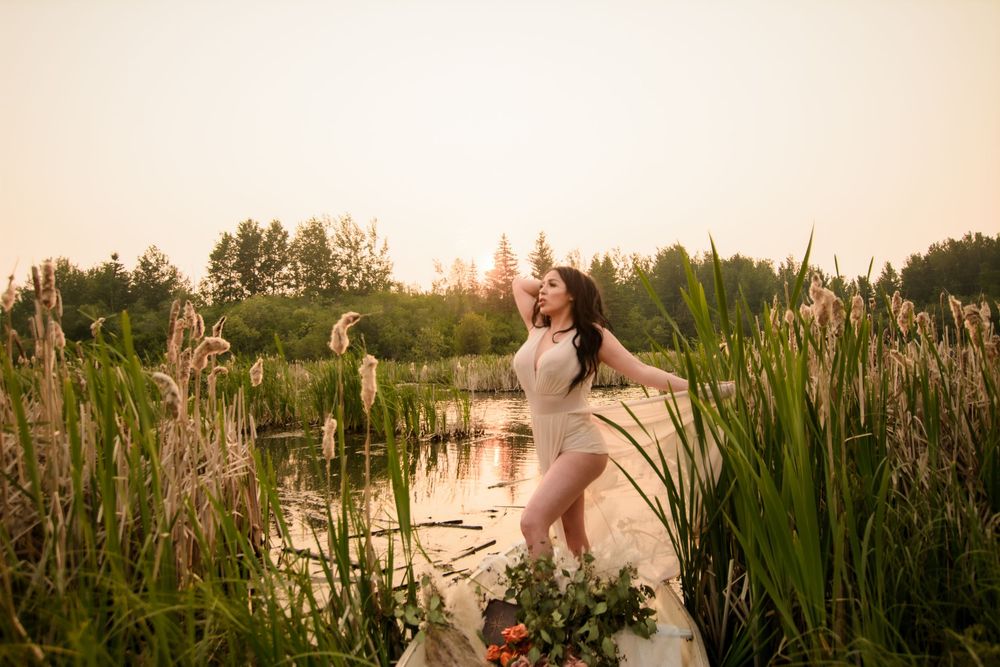 Ballet dancer poses gracefully among cattails at sunset by a serene pond while wearing a white leotard.