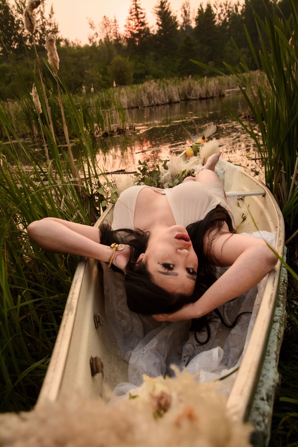 A white bathtub sits nestled among tall reeds and grass at sunset near a lake.